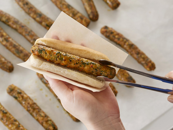 Tongs placing a veggie sausage into a hot dog bun held in a hand.