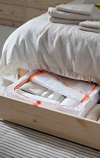 Wooden underbed drawer with a white storage bag featuring an orange zipper, used for storing bedding or textiles, under a bed dressed in light-colored blankets.
