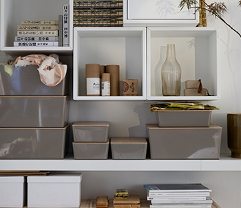 White shelving unit with glossy grey storage boxes, decorative items like glass bottles, books, and cylindrical containers, in a modern and neatly organized space.