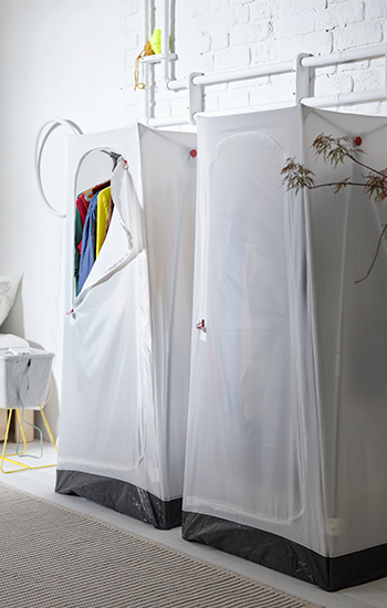 White portable wardrobes with tubular frame and semi-transparent fabric cover, one open showing hanging clothes on hangers, in a bright room with a white wall.