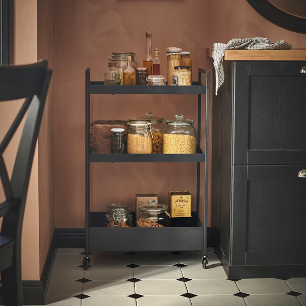 A black three-tier utility cart with food jars, next to a dark cabinet and pink wall.