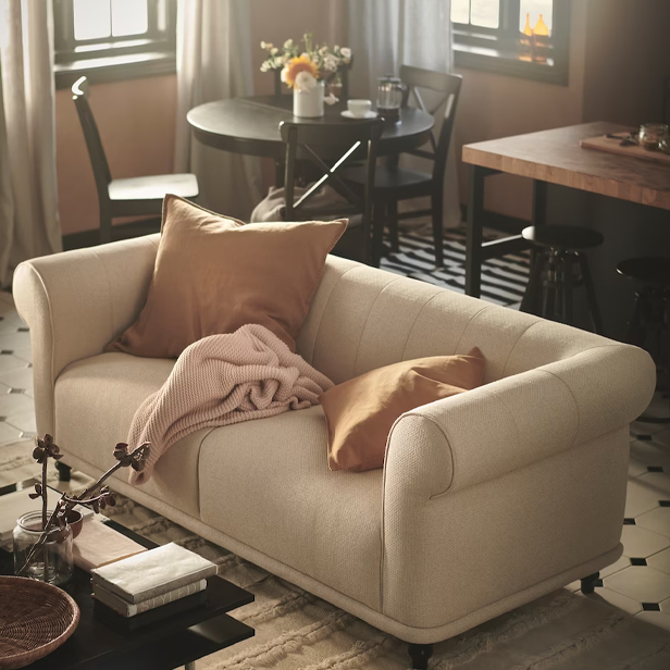A beige sofa with brown cushions and a pink blanket, in a room with a dining and kitchen area in the background.