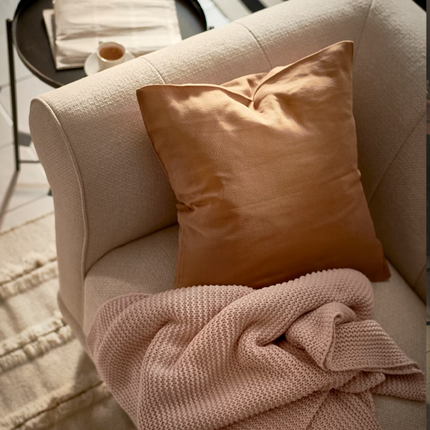 A beige single sofa with a brown cushion and a pink knitted blanket, with a side table in the background.