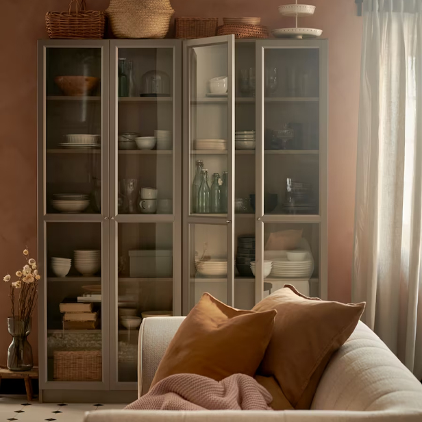 A pair of gray display cabinets showcase dinnerware and glassware in a warm living room. Ochre cushions in the foreground.