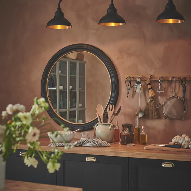 A rustic kitchen with a pink wall, round mirror, black cabinets, and a wooden countertop.