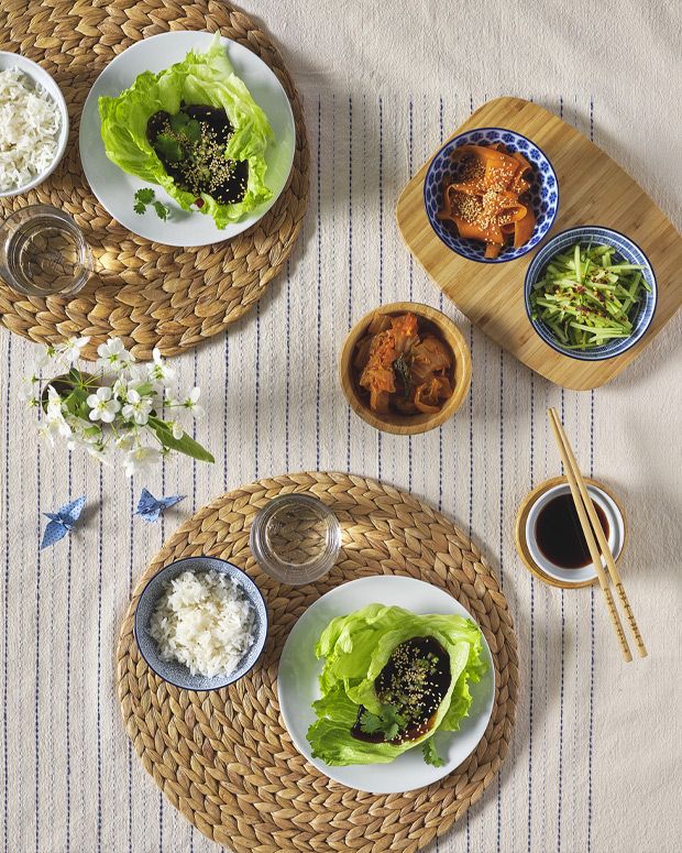 A cozy table setting with rice, lettuce wraps, and side dishes, served with chopsticks on woven placemats.