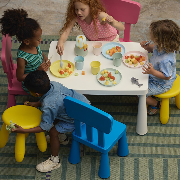 Children seated around a small white table with pastel colored chairs, served pastel colored plates served there are squared fruits