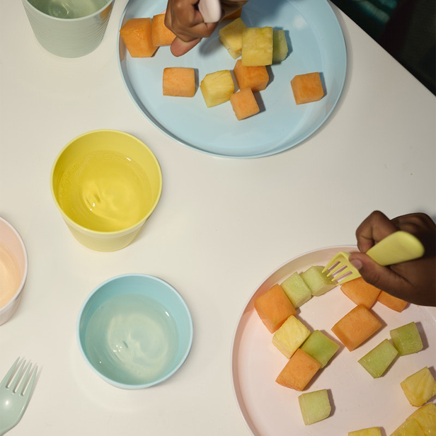 Pastel-colored plates, with fruit cut into squares, with children's hands eating.