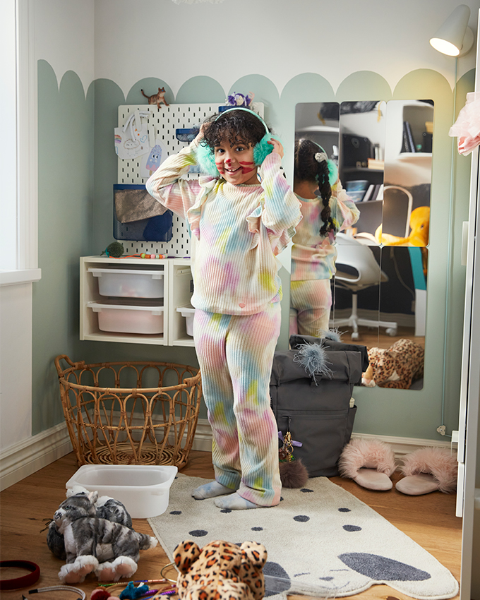 A smiling girl in furry earmuffs and pastel pajamas in a messy, playful room.