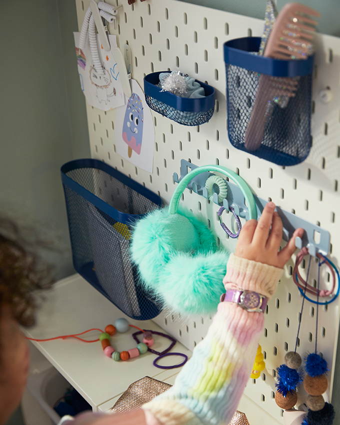 Child's hands organizing a white pegboard with blue baskets, mint earmuffs, and colorful jewelry.