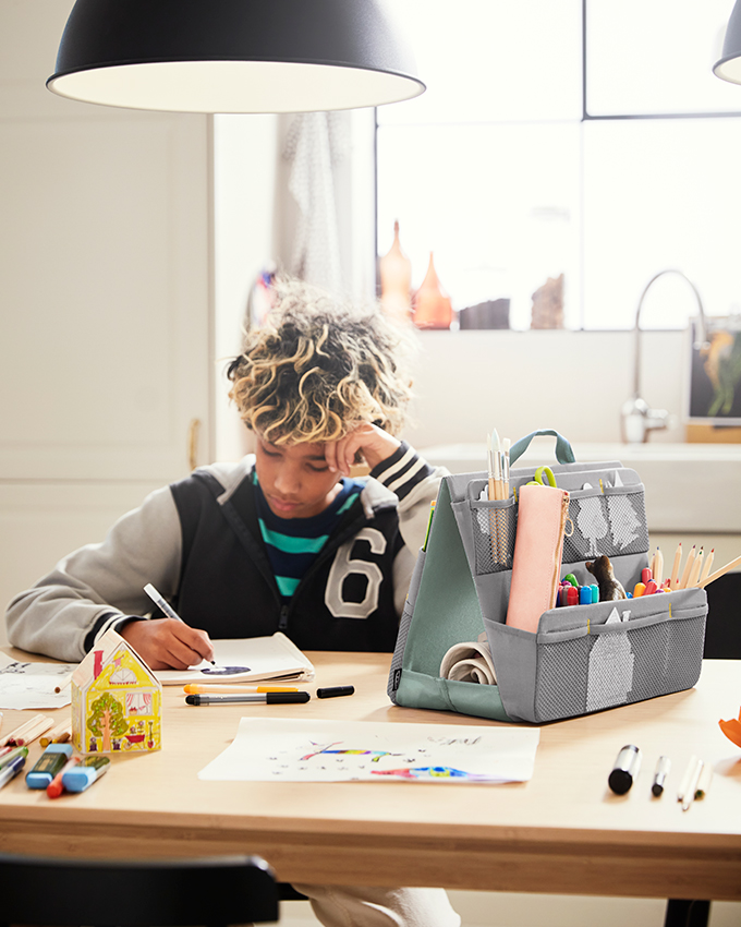 A child drawing intently at a wooden table with a gray art organizer and colorful accessories.