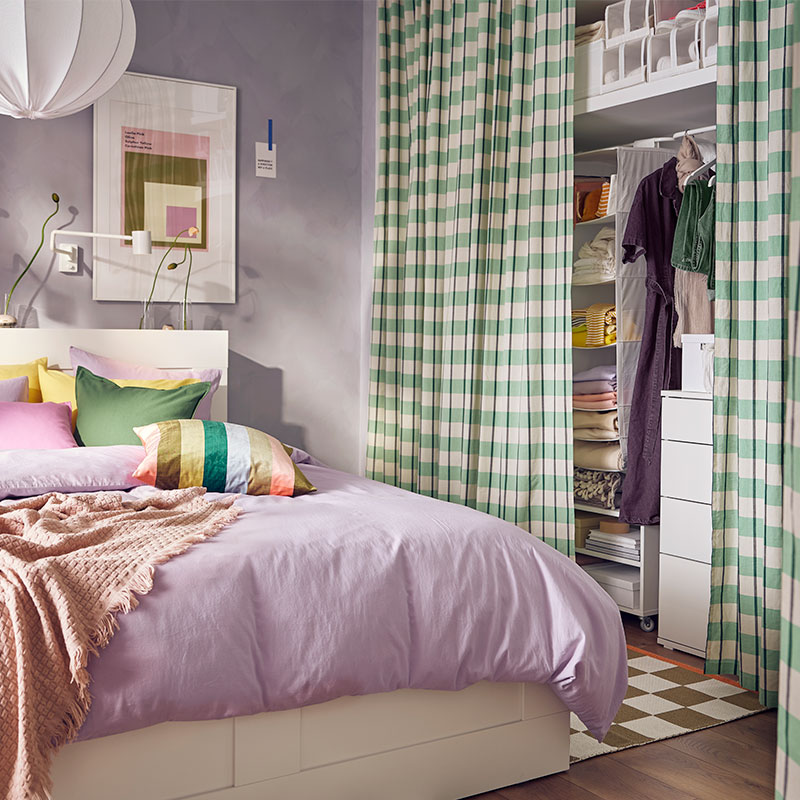 Colorful bedroom, a large bed with brightly colored cushions, a light purple cover and a closet with green plaid colors.