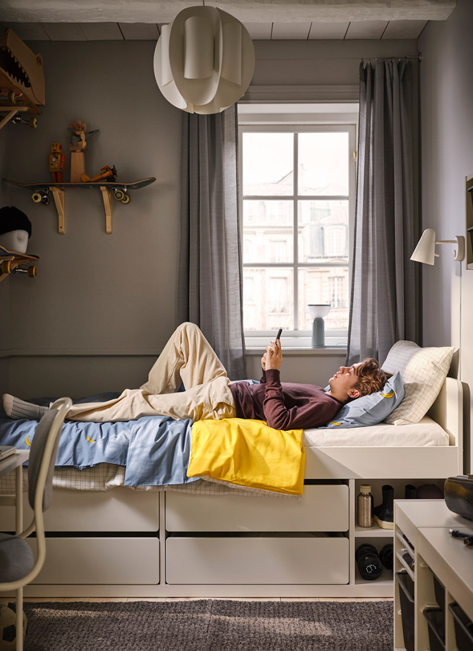 A youth room with gray walls, a bed with drawers, blue sheets, and a yellow blanket. A young person uses their phone.
