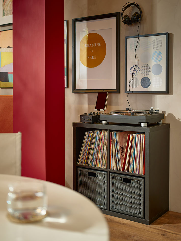 open storage cabinet with four spaces, with wicker boxes in the lower one and full of vinyl records on top, there is a record player on the top, and on the wall pictures of records.
