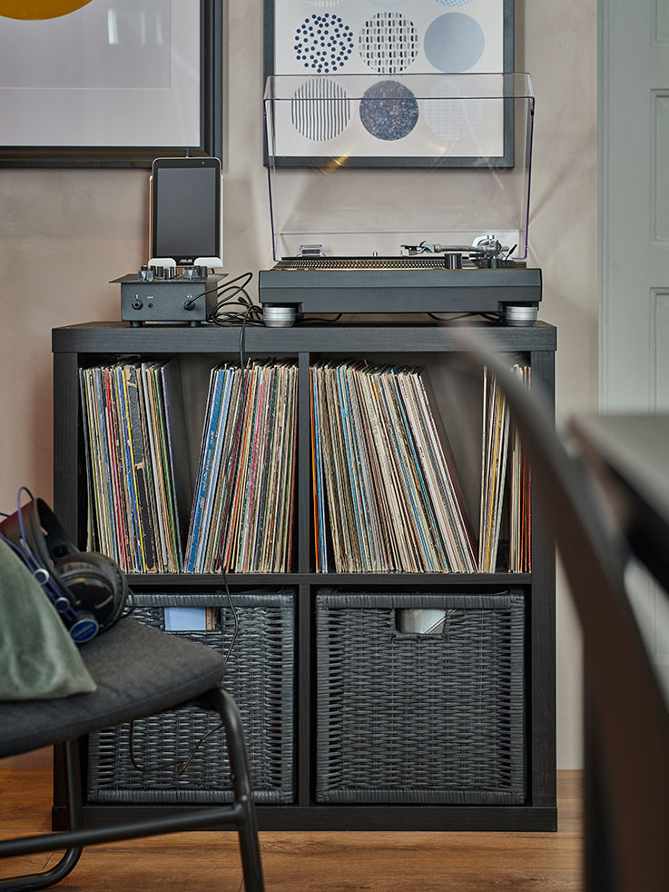 open storage cabinet with four spaces, with wicker boxes in the lower one and full of vinyl records on top, there is a record player on the top, 