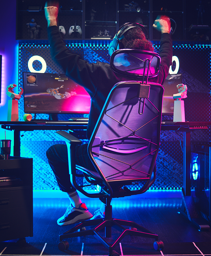 A gamer celebrates victory in a room lit by vibrant blue and pink neon lights. They sit on a black mesh chair in front of a gaming PC with a glowing "O" monitor.