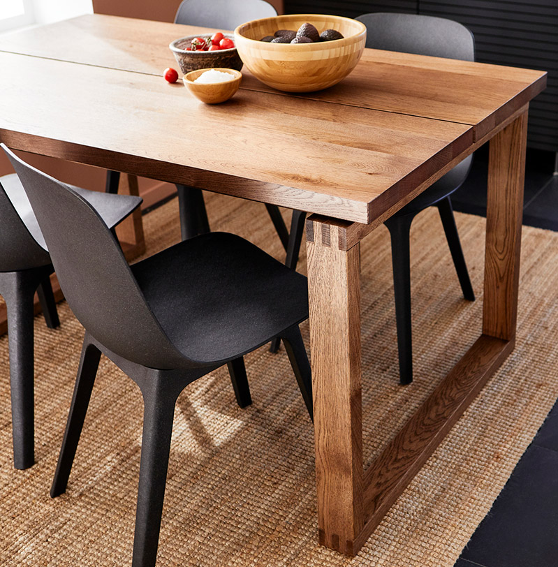 Wood dining table MÖRBYLÅNGA with modern black chairs on woven rug, minimalist Scandinavian style.