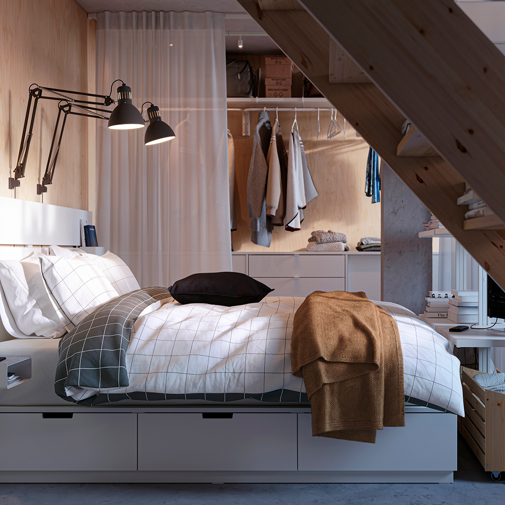Compact bedroom with white bed, built-in storage, and exposed wooden staircase.