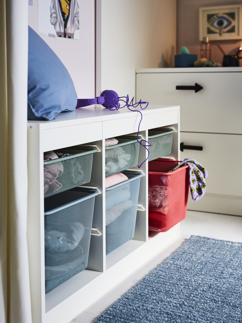 A white bench with mesh baskets in pastel and red tones, on a blue rug. An organized and functional ambiance for storing clothes or toys.