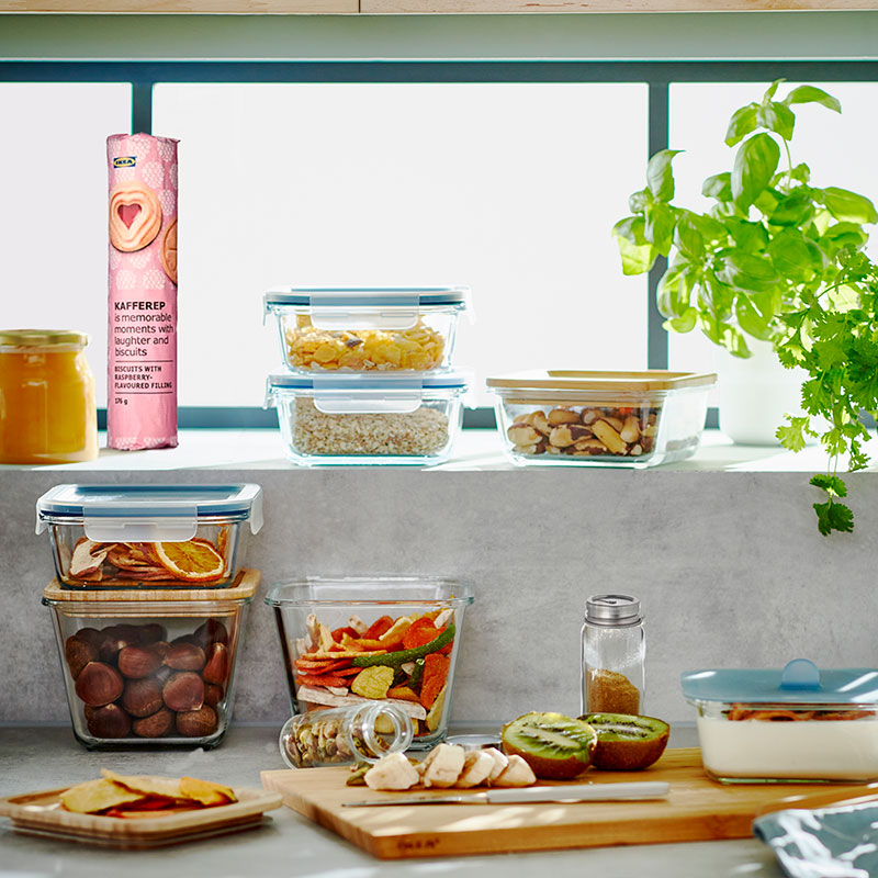 Rustic outdoor kitchen with food containers, sliced fruits, preserves, and a cutting board with knife in a cozy, natural setting.