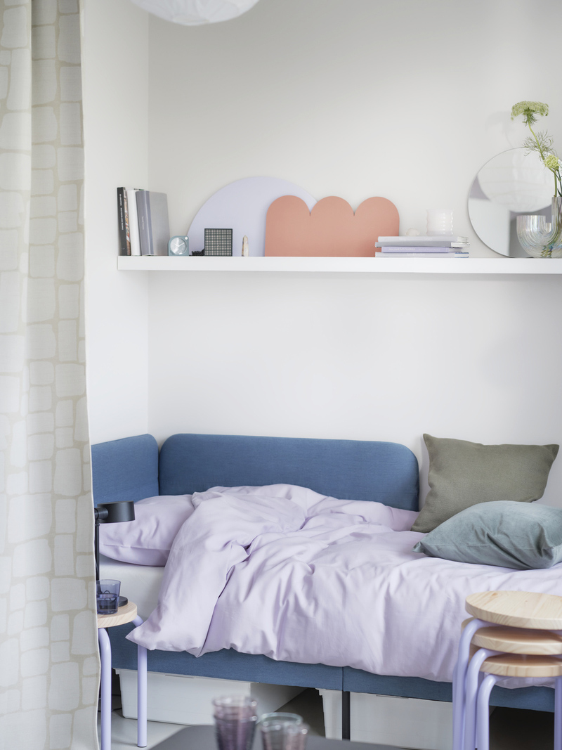 Studio apartment with a bed next to several stacked DOMSTEN stools, featuring a LACK shelving unit with a BLÅSER mirror and a RÄFFELBJÖRK vase.