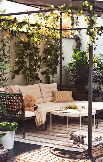 Rincón exterior con sofá rinconero de ratán con cojines beige, decorado con plantas trepadoras, peluches y una mesa redonda blanca, bajo una pérgola adornada con guirnaldas de hojas verdes.