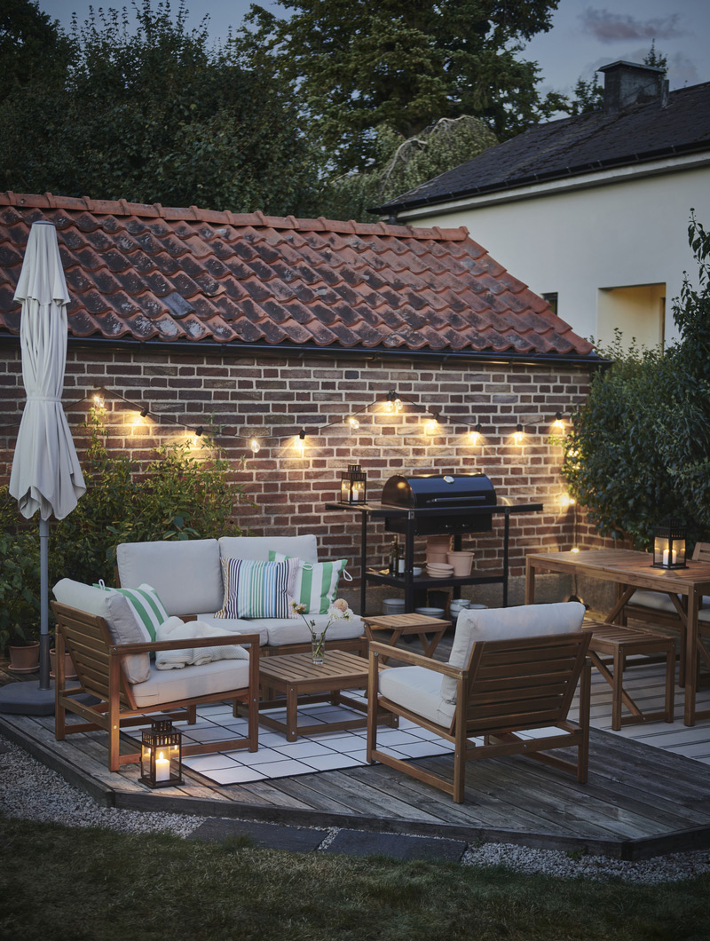 Patio iluminado con luces cálidas, muebles de madera clara y cojines verdes y blancos. Un ambiente acogedor para las noches al aire libre.