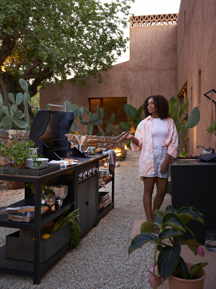 Mujer cocinando en una parrilla exterior en un patio con plantas y paredes color tierra. Un ambiente cálido y relajado.