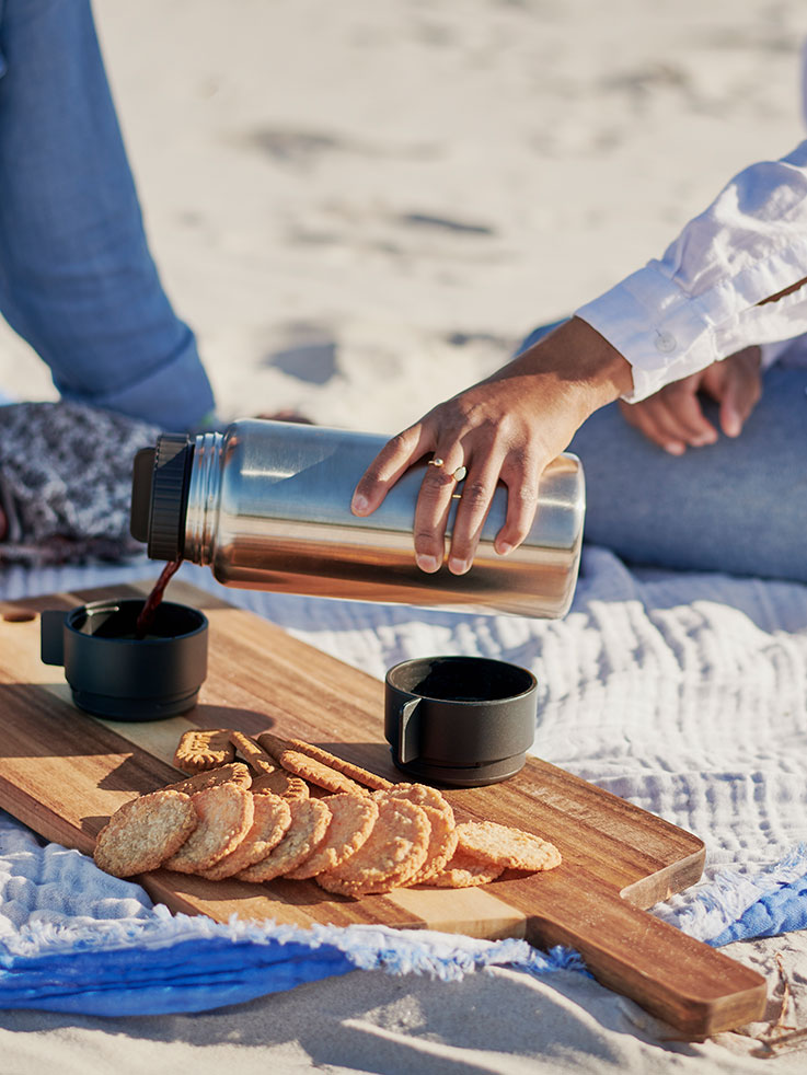 Person pouring coffee from a thermos on the beach, with cookies on a wooden board.