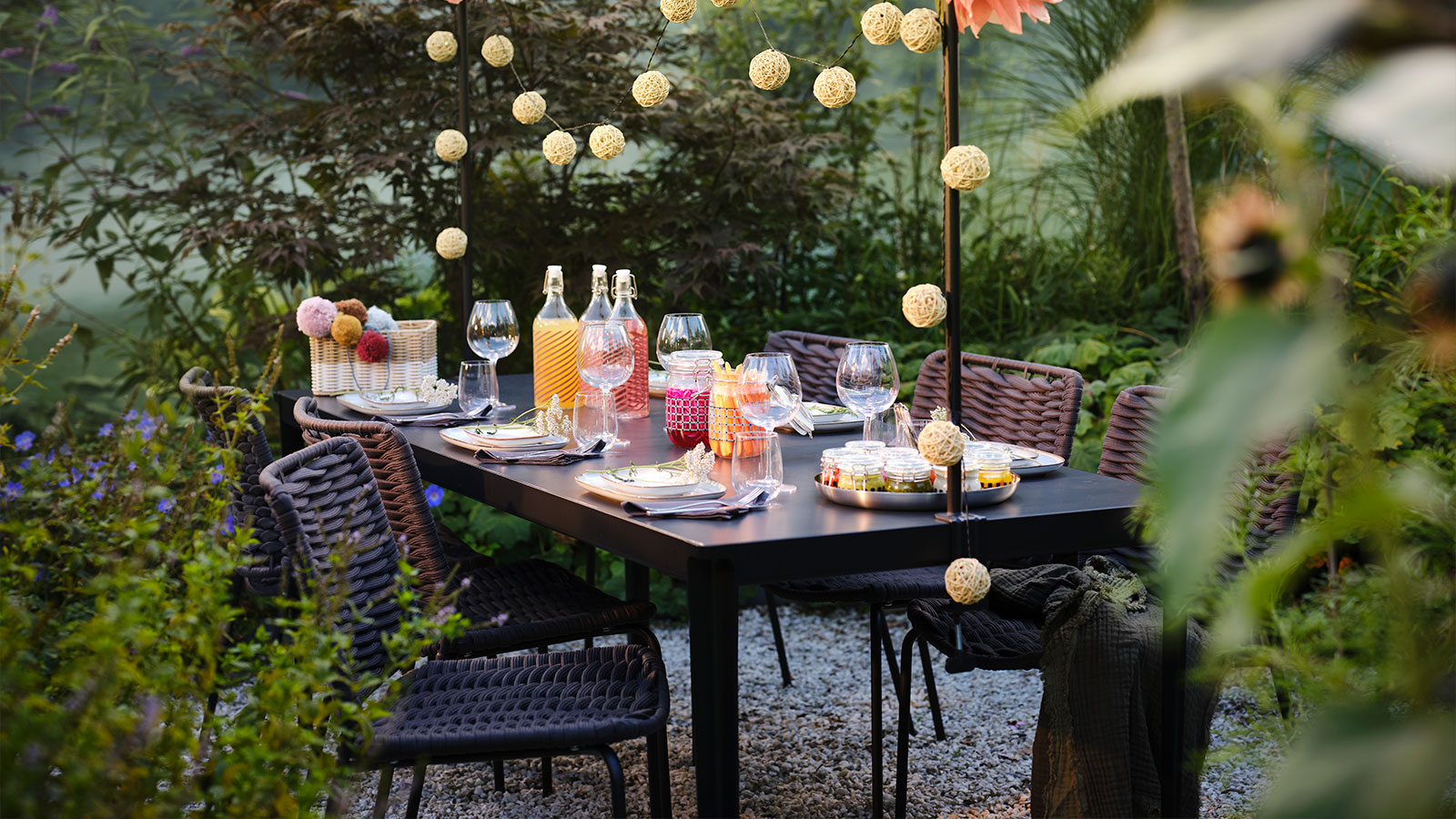 Outdoor table set with drinks and decorations, hanging lights. Festive and natural ambiance.