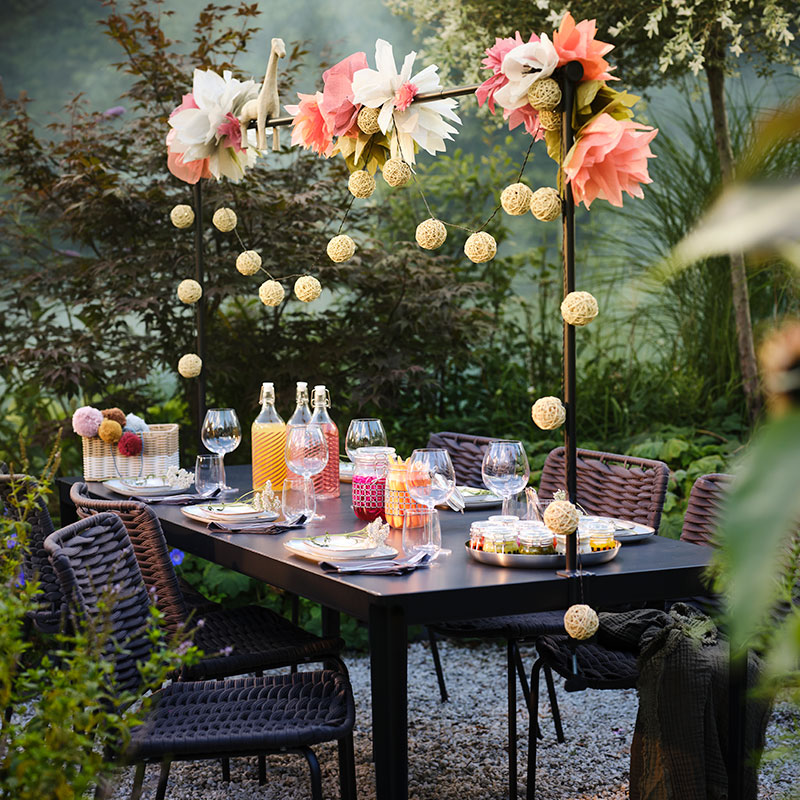 Outdoor table set with drinks and decorations, hanging lights. Festive and natural ambiance.