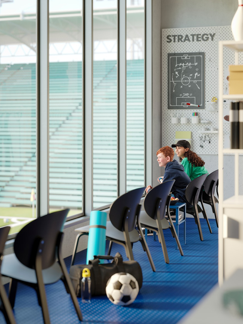 A waiting room with modern dark-colored armchairs, a soccer ball on the floor on a blue carpet, and a large window with a view of a sports field.