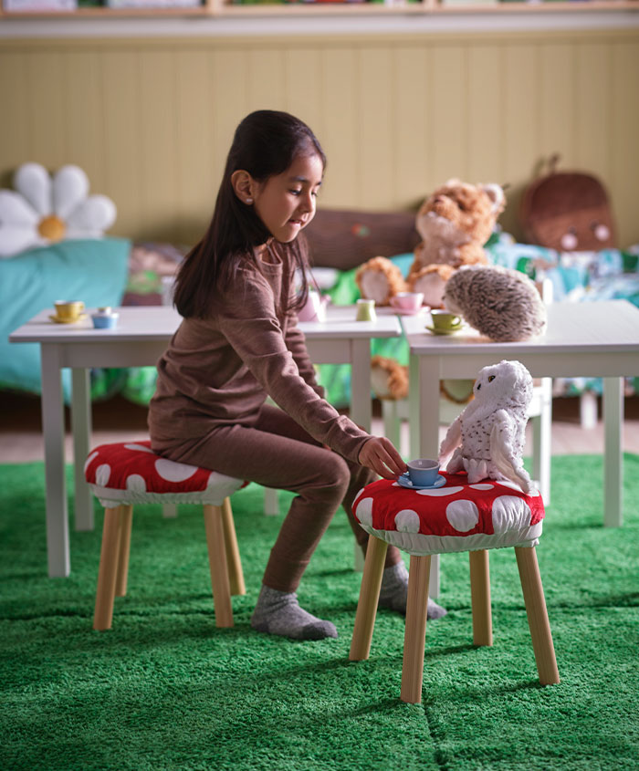 Girl in brown pajamas sitting at a tea table with mushroom stools and plush toys. Playful atmosphere.