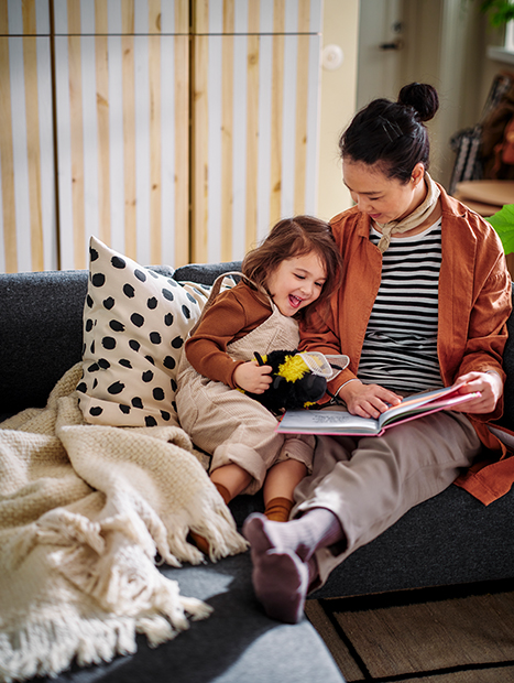 Madre e hija leyendo juntas en el sofá, con manta y cojín decorativo en un ambiente acogedor.
