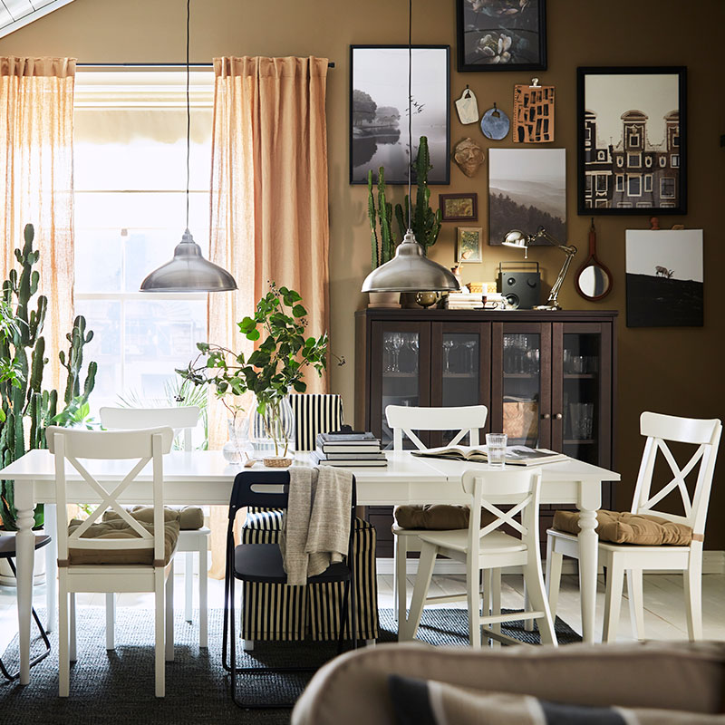 Traditional style dining room with vintage touches, white table, chairs with cushions, a large window with beige curtains, on the wall to the right a bookshelf and display case, in the background a showcase.