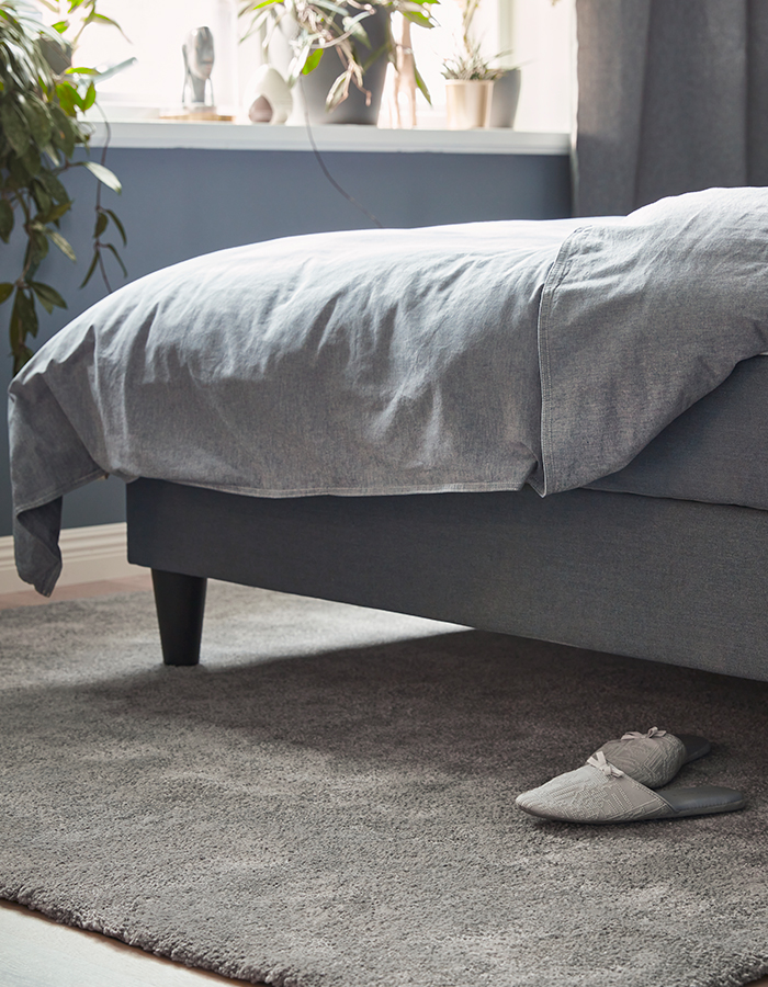 Grey upholstered bed with matching duvet, placed on a soft rug, with grey slippers beside the bed and decorative plants on the windowsill.