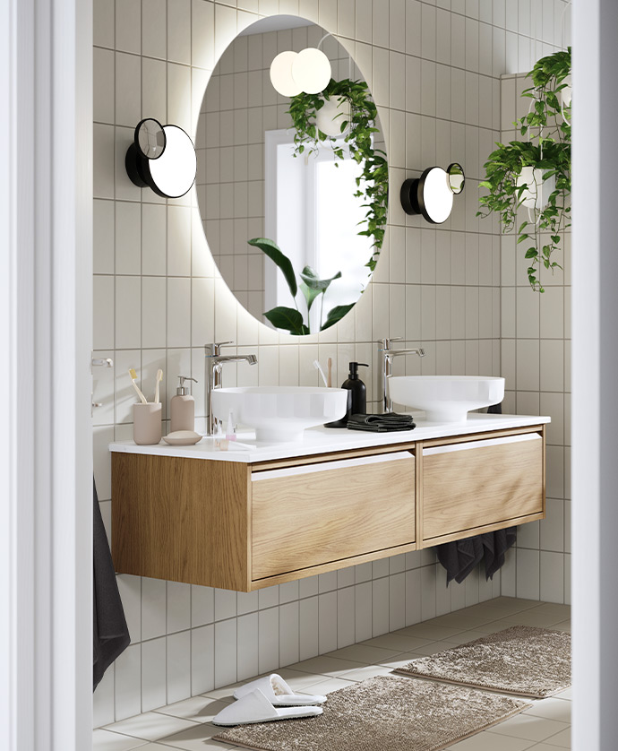 Baño moderno con mueble flotante de madera, dos lavabos redondos blancos, espejo ovalado iluminado y apliques negros, decorado con plantas colgantes y alfombrillas beige.