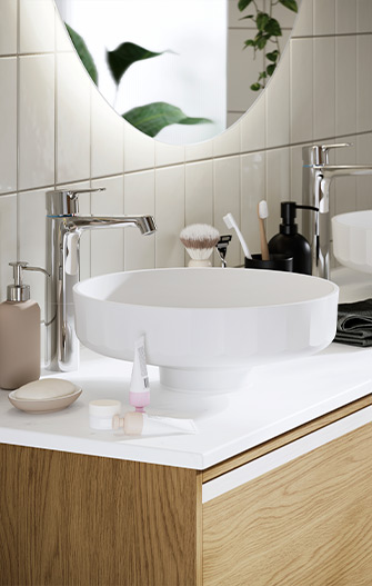 Primer plano de lavabo redondo blanco sobre encimera blanca con grifería cromada, mueble de baño de madera y accesorios de aseo como jabón, brocha y dispensadores.