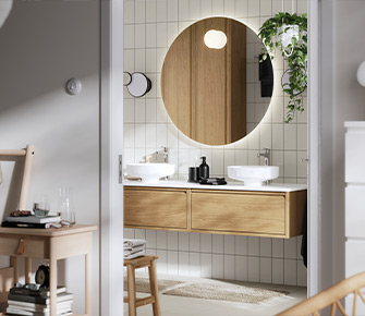 Vista desde el dormitorio hacia un baño moderno con mueble flotante de madera, dos lavabos blancos, espejo redondo iluminado y plantas colgantes sobre pared de azulejos.