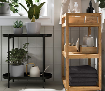 Soporte para plantas negro junto a un carrito de bambú con productos de cuidado personal en un baño de azulejos blancos. 