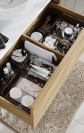 Cajón de muebles de baño abierto de color marrón claro con productos de aseo bien organizados en cajas de plástico; hay una alfombra de baño en el suelo.
