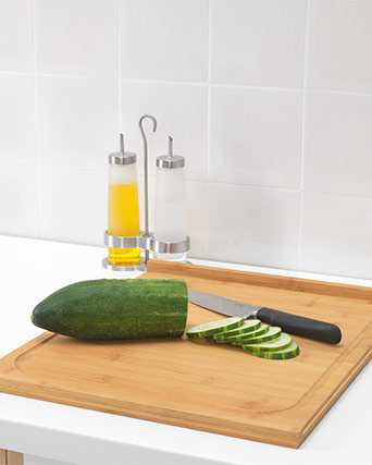 A half-sliced green cucumber and a black-handled knife rest on a light brown bamboo cutting board. Stainless steel oil and vinegar dispensers stand nearby.