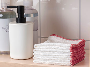 White soap dispenser with black pump, two other bottles, and a stack of white and red-edged towels on a light wooden surface.