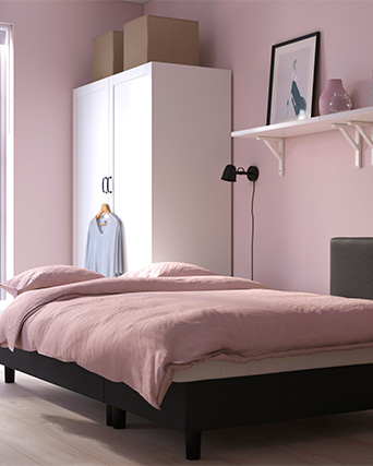 A cozy bedroom with pink walls, a neatly made bed, a white wardrobe, and decorative items on a shelf.