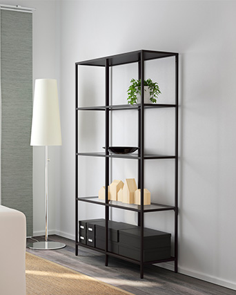 Una estantería alta de metal negro contiene una planta verde, un cuenco negro, modelos de casas de madera y cajas negras. Lámpara de pie blanca a la izquierda.