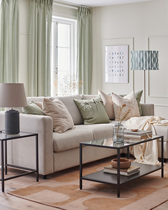 Sofá beige, cojines variados, mesas oscuras, lámpara negra, lámpara de pie con patrón azul. Cortinas verde claro y decoración.