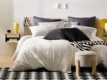 IKEA bedroom decorated with light and dark textiles, a rug, and a yellow table lamp.