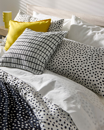 Neatly made bed with several patterned pillows (polka dots and grid designs), featuring a yellow pillow. The bedding includes a white sheet and a polka dot comforter. In the background, a bedside table holds a yellow cup or container.