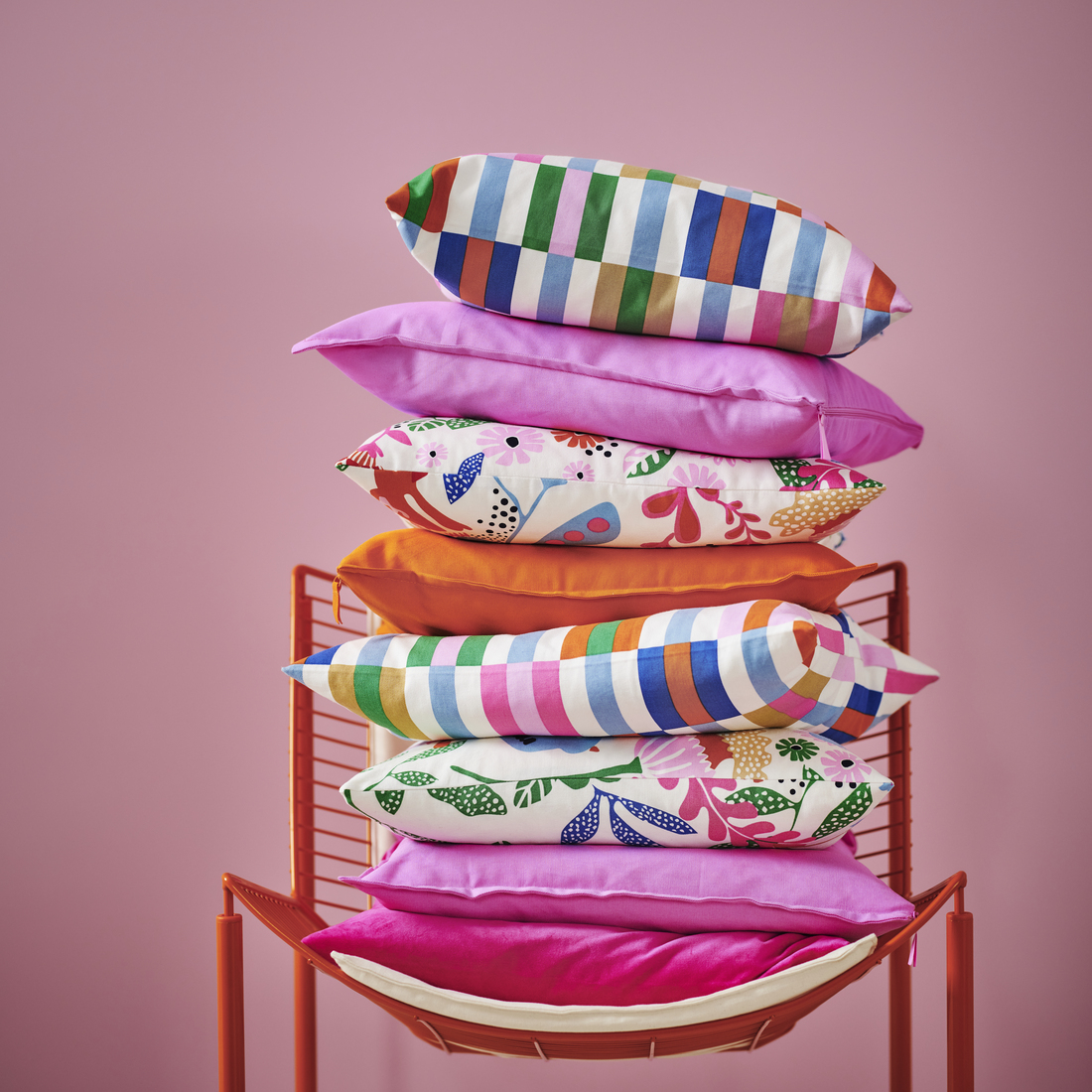 Stack of colorful and patterned cushions on an orange metal chair against a pink wall.