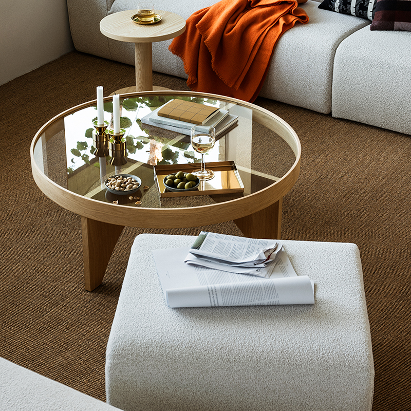 A cozy living room with a glass coffee table, snacks, a drink, and soft white furniture accented by a bright orange blanket.